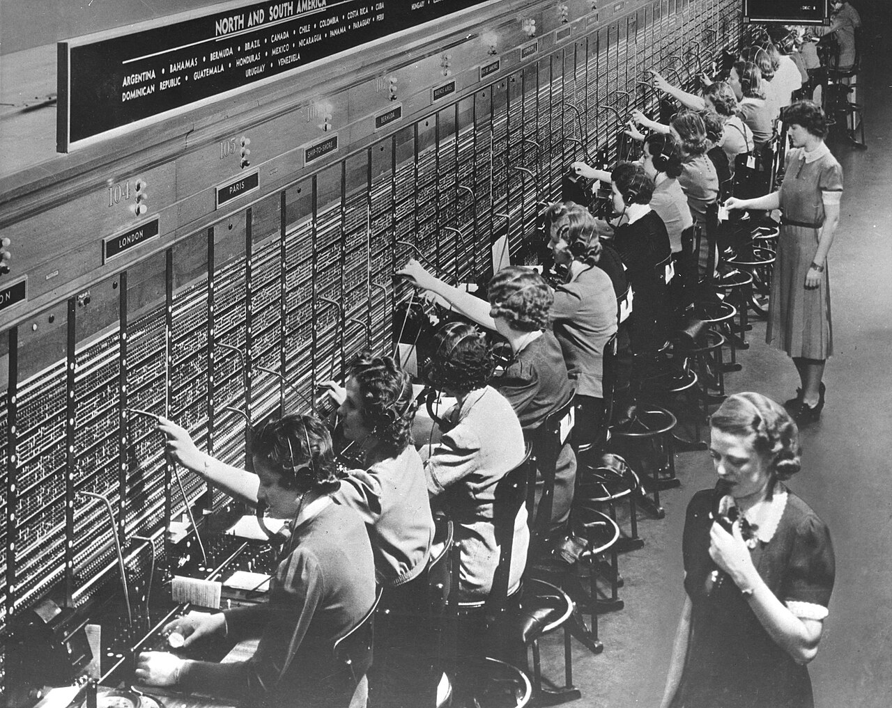 A long row of telephone operators in headsets plugging cords into a tall switchboard labelled London and Paris.